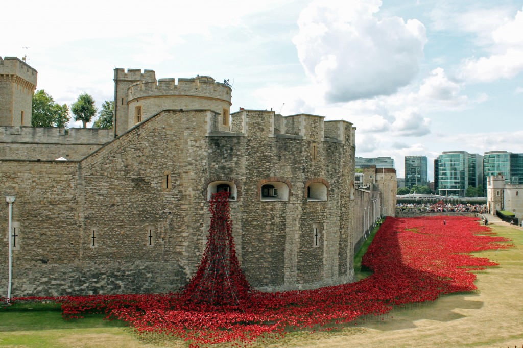 Image of the exterior walls of the Tower, with an art installation that features red metal poppies which appear to pour out of one of the windows of the wall and spread along the grassy floor of the moat.