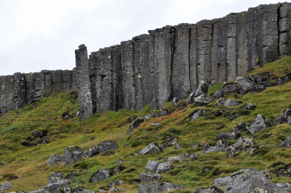 Dark grey basalt columns rise high above a bright green field strewn with boulders