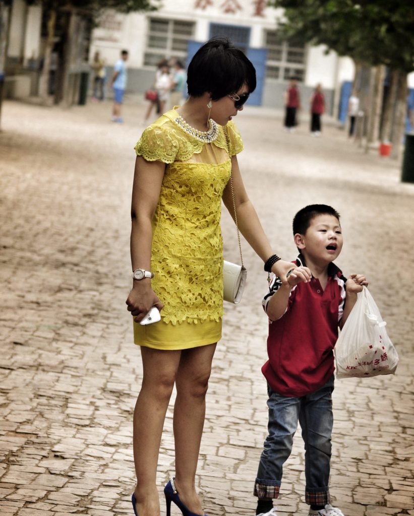 A mother in a fashionable yellow dress holds the hand of a young boy in uniform. He looks unhappy.
