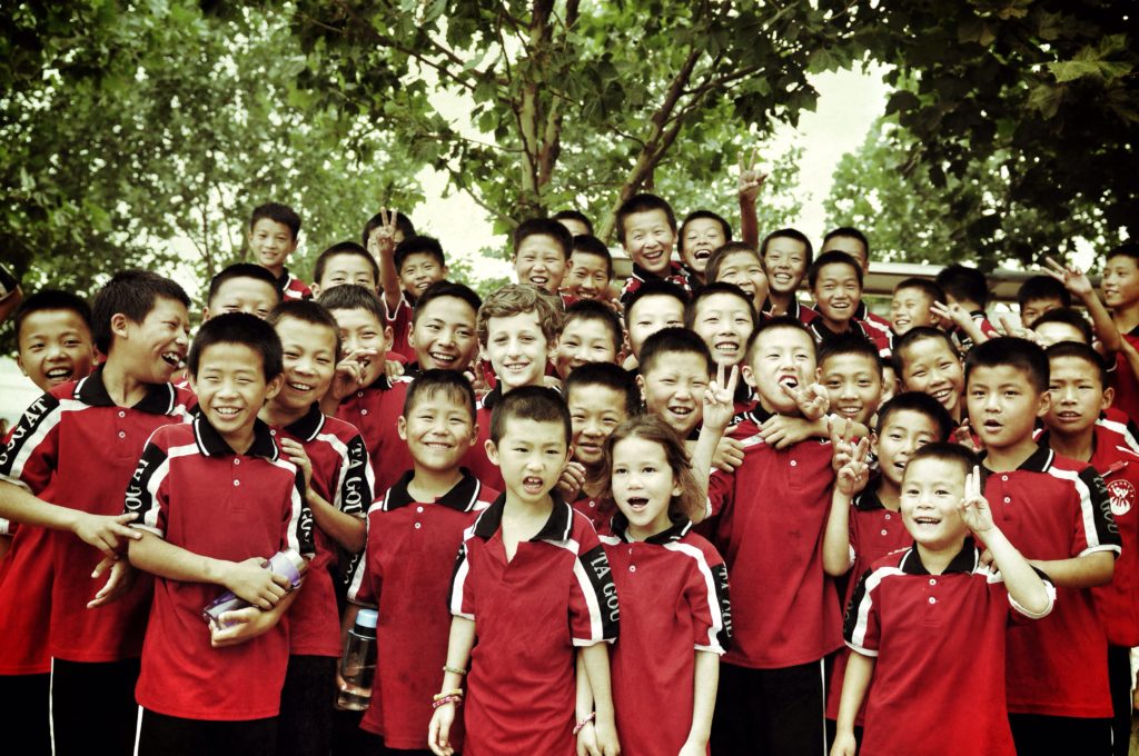 A large group of children, Chinese and Caucasian, wearing the uniform of the monastery, pose smiling for the camera.