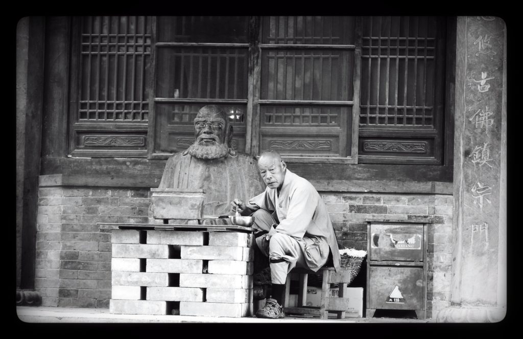 An elderly man sits at a makeshift table of bricks eating noodles in front of a statue of Buddha.