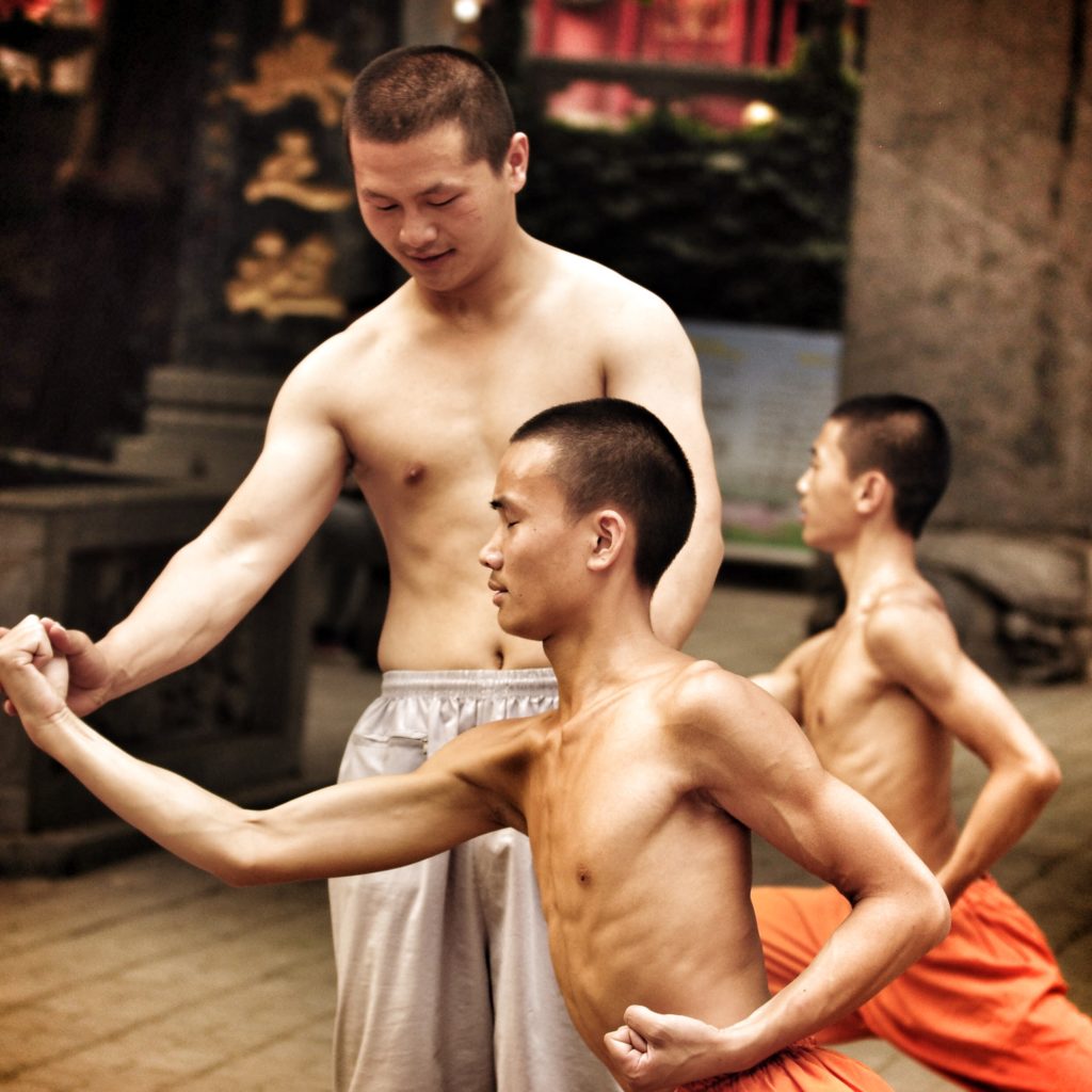 An instructor adjusts the stance of a young man practicing a wushu form, eyes closed.