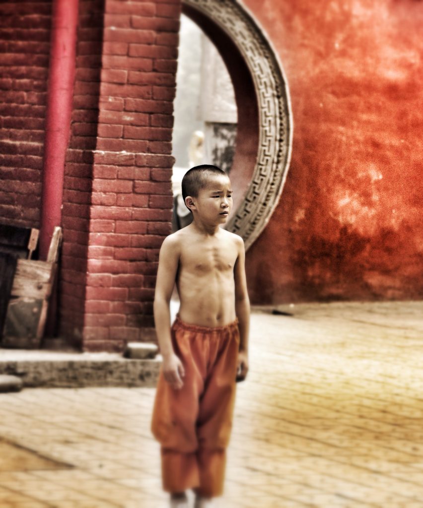 A young boy stands in front of a Chinese key-shaped door wearing the orange pants of a Shaolin warrior. 