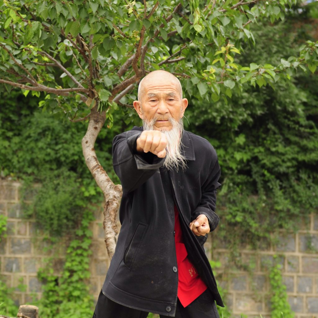 And elderly man with a long white beard extends a fist toward the camera in a wushu form.